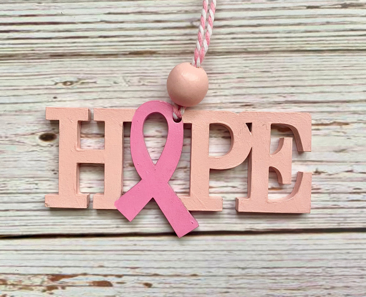 A wooden HOPE hanging ornament with a pink ribbon over the letter O, hanging against a whitewashed wood plank background.