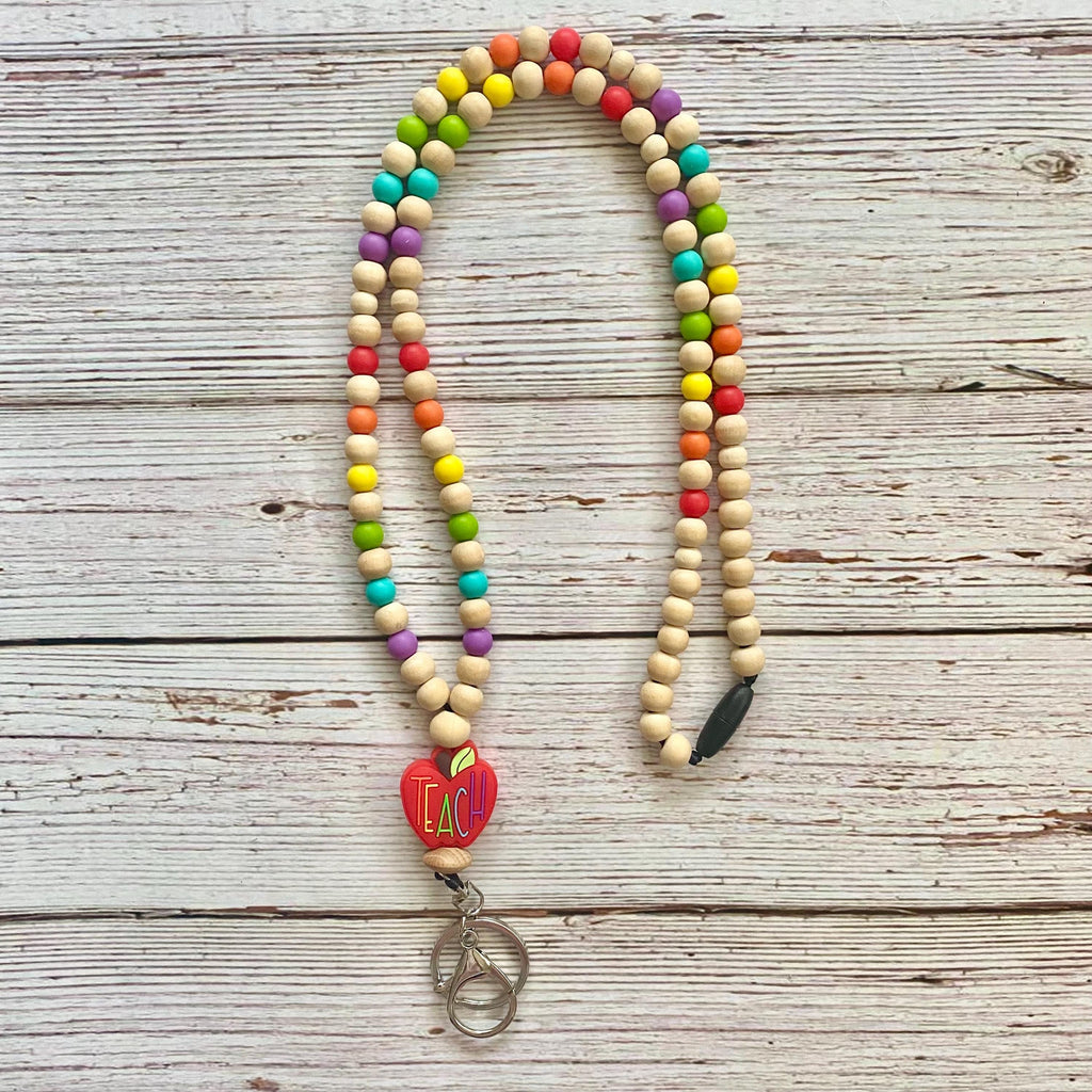A beaded necklace with natural wood beads and colorful accent beads, featuring a red heart charm with 'TEACH' and silver key rings, laid flat on a rustic white-washed wooden background.
