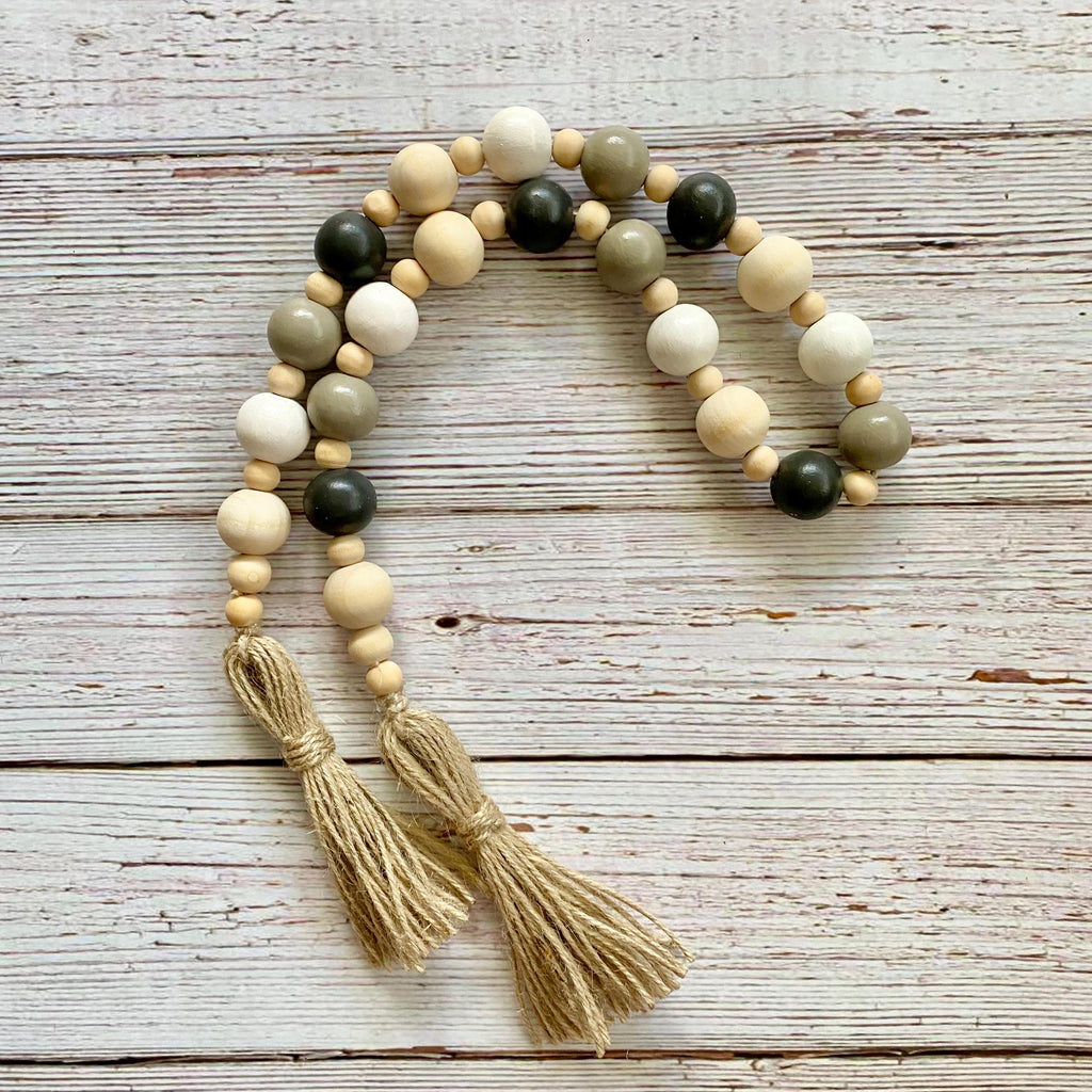 A beaded garland with natural wood beads in cream, tan, olive green, and black, featuring two knotted jute tassels, displayed on a rustic white-washed wooden background.