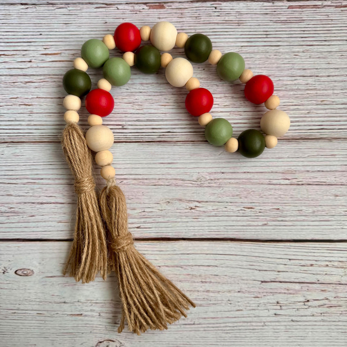 A beaded garland with alternating red, sage green, olive green, and natural wood beads, finished with two long tan jute tassels, laid flat on a whitewashed wooden surface.