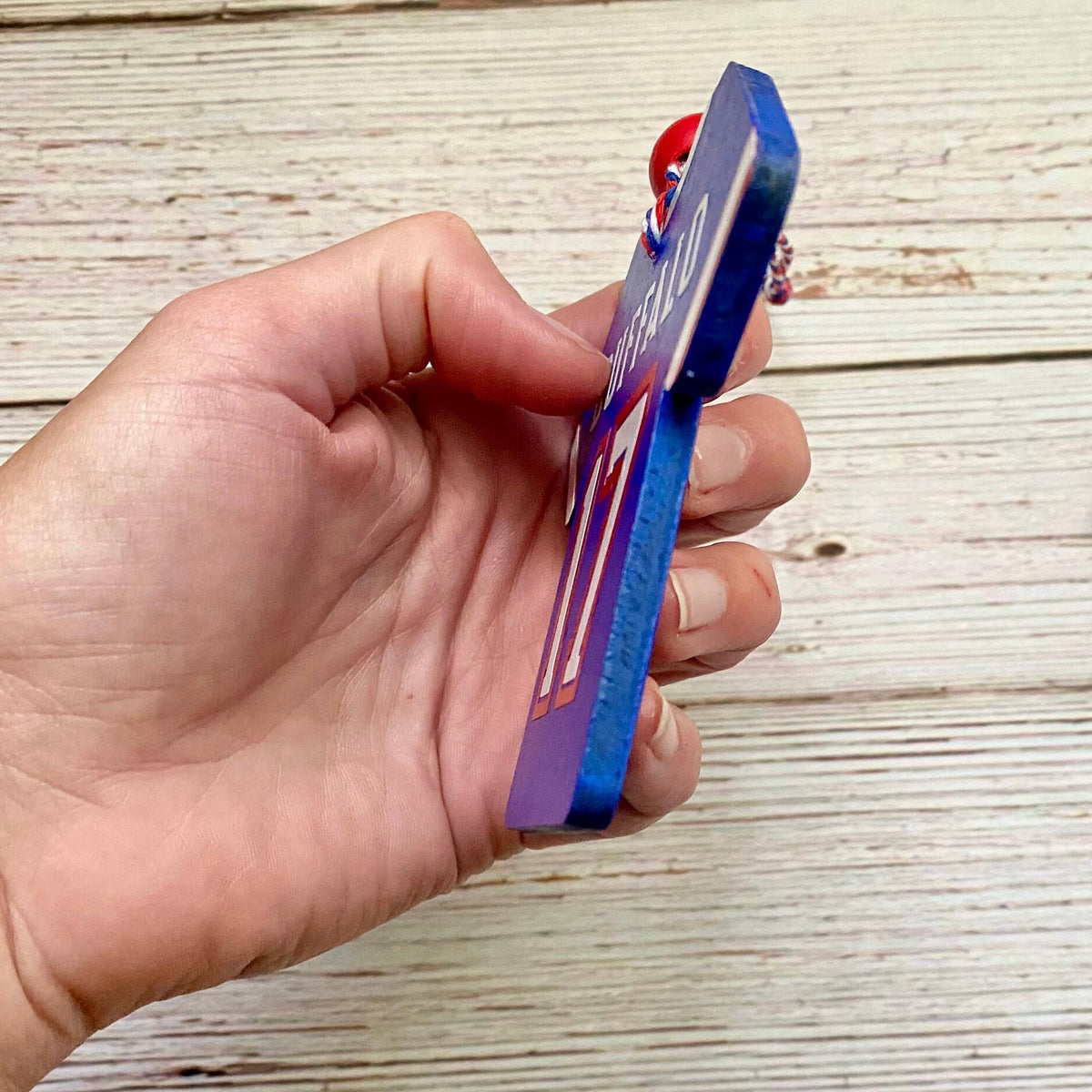 A hand holding a blue wooden name plaque or keepsake box with a red and white beaded keychain, shown against a light wood plank background.