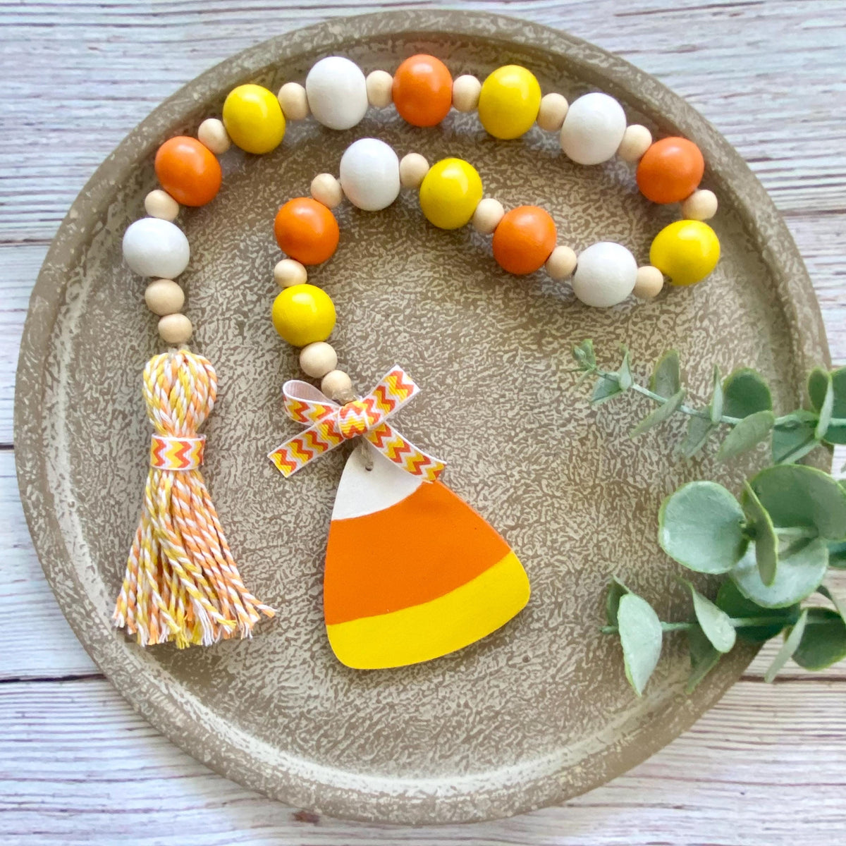 A beaded garland with orange, yellow, and white wooden beads, a tassel, and a candy corn charm, displayed on a round textured tray with eucalyptus leaves.