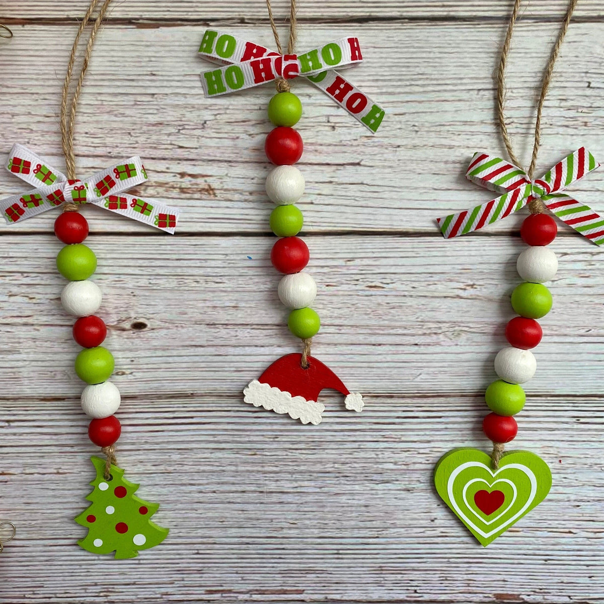 Three handmade Christmas bead garlands hanging on a whitewashed wooden surface. The garlands feature red and green beads, white beads, jute hangers with patterned paper bows, and small holiday decorations including a green Christmas tree, a red Santa hat, and a green heart with a red heart.