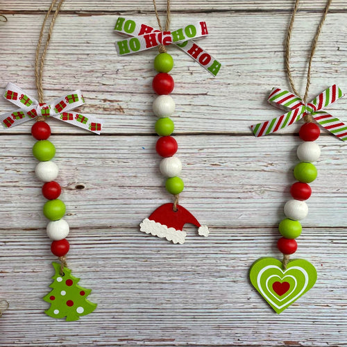 Three handmade Christmas bead garlands hanging on a whitewashed wooden surface. The garlands feature red and green beads, white beads, jute hangers with patterned paper bows, and small holiday decorations including a green Christmas tree, a red Santa hat, and a green heart with a red heart.