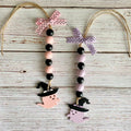 Two Halloween bead garlands with black and pastel pink wooden beads, witch hats, ghost cutouts, and ribbon hangers on a whitewashed wood background.