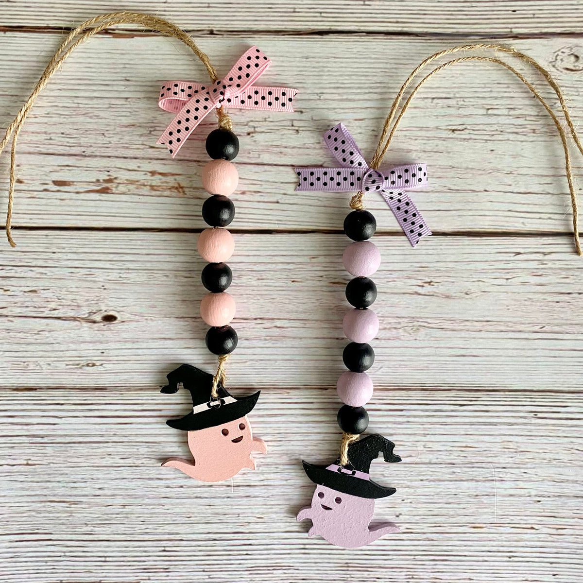 Two Halloween bead garlands with black and pastel pink wooden beads, witch hats, ghost cutouts, and ribbon hangers on a whitewashed wood background.