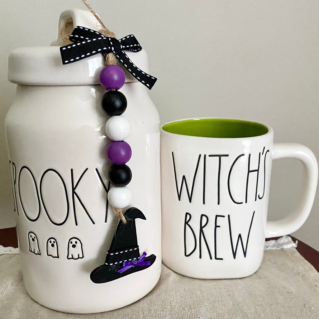 A white ceramic Halloween-themed canister with a lid and a matching white mug with a green interior. The canister is printed with the word 'BOOKEY' and small ghost icons, decorated with a black-and-white ribbon, purple, black, and white bead garland, and a black witch hat.