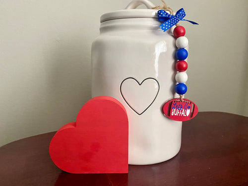 A white ceramic jar with a lid, decorated with a black heart outline, a blue polka-dot ribbon, and a red, white, and blue beaded charm with a red 'Buffalo' tag. A red wooden heart sits on a dark wooden table in front of the jar against a light beige wall.