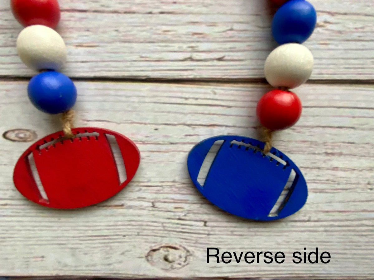 A close-up of a beaded garland laid on a whitewashed wood surface, showing red and blue football-shaped charms with cutout details. The beads are red, white, and blue, and the text 'Reverse side' appears at the bottom right.