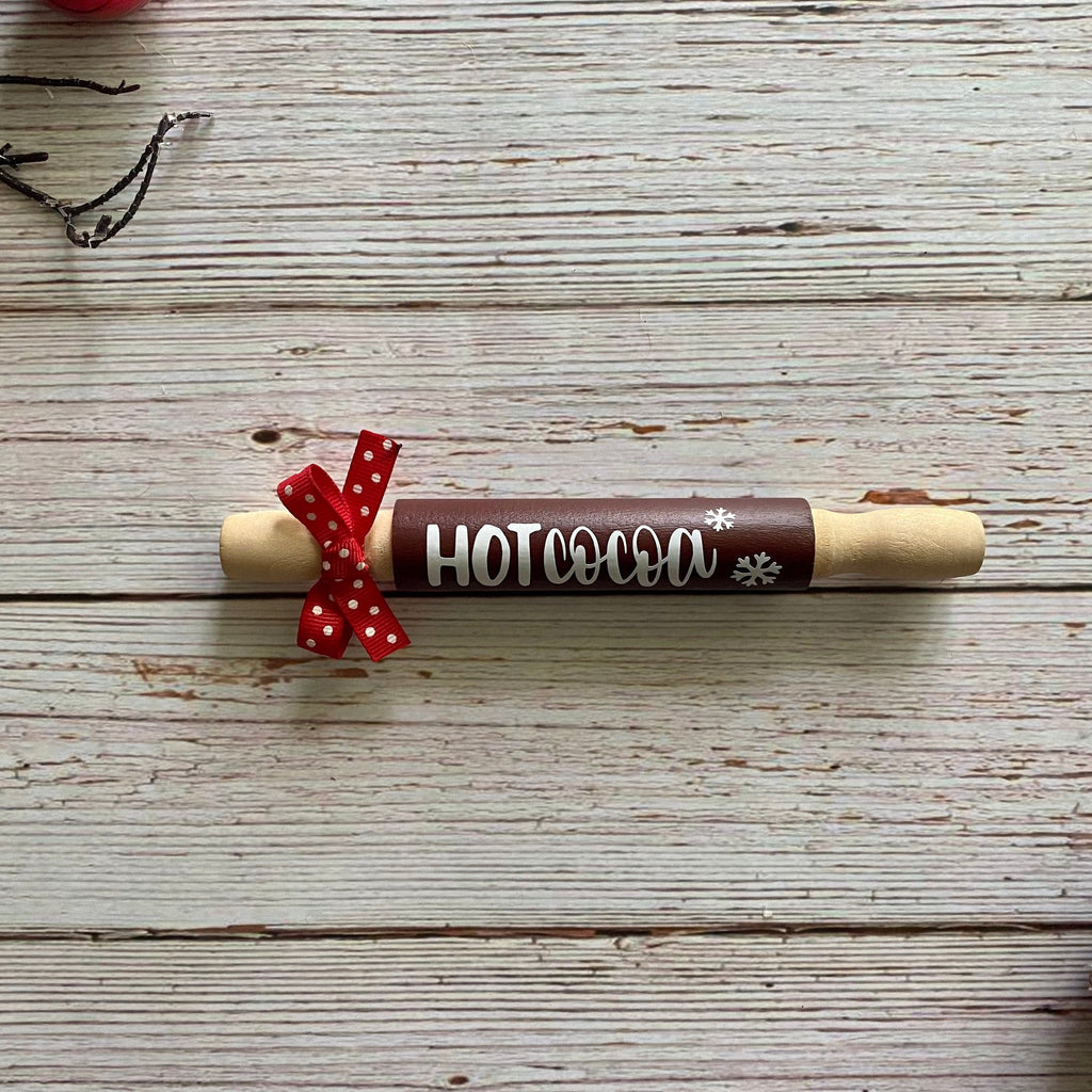 A wooden hot cocoa holder shaped like a rolling pin, featuring a dark brown center with the words 'HOT cocoa' in white and a red polka-dot ribbon tied around it, displayed on a rustic white wood plank background.