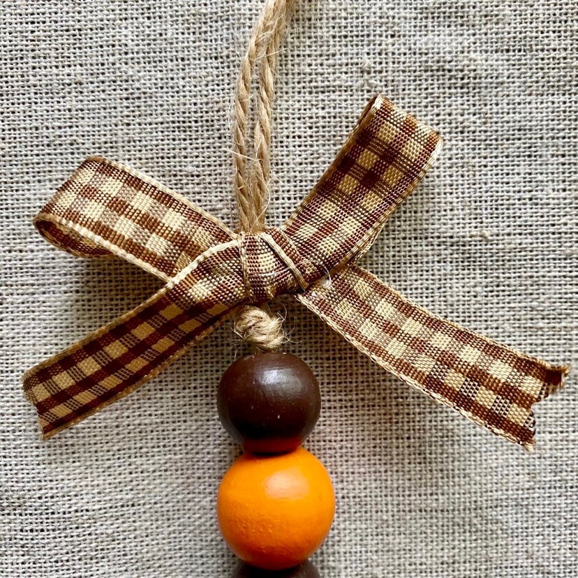 A rustic Christmas ornament featuring a brown and beige gingham ribbon bow, a natural jute hanging loop, and two wooden beads, one dark brown and one orange, set against a textured beige burlap background.