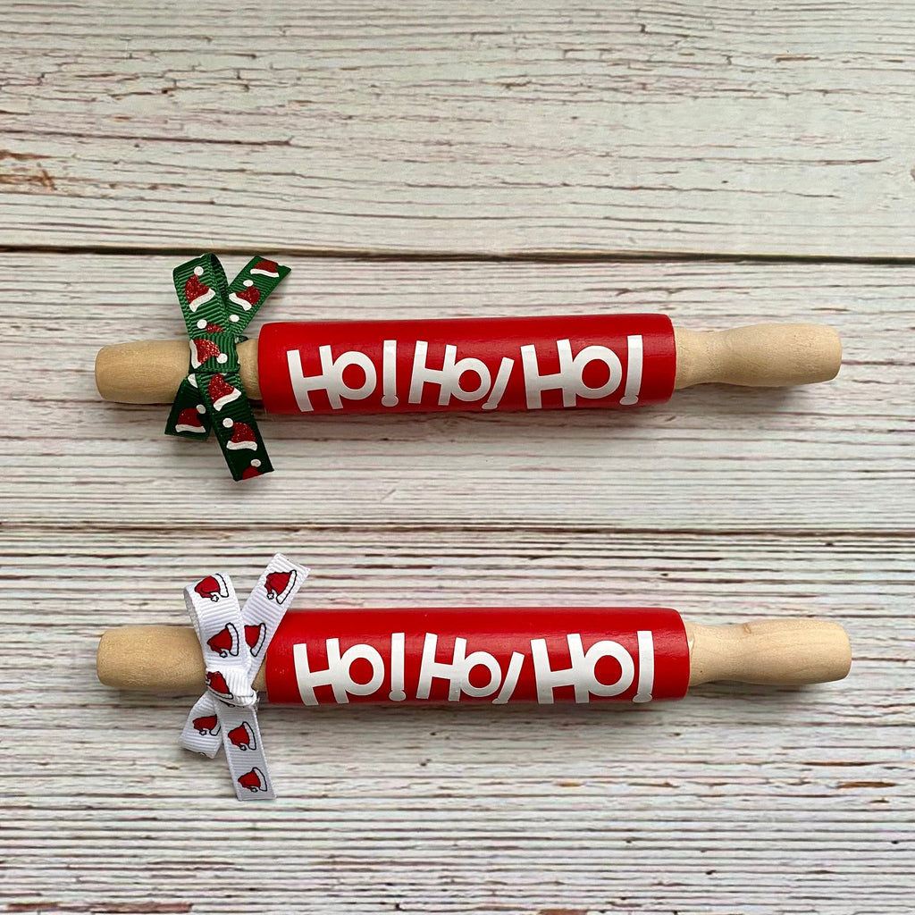Two wooden rolling pins with red handles printed with 'Ho Ho Ho' in white, tied with festive ribbon bows, displayed on a whitewashed wooden background.
