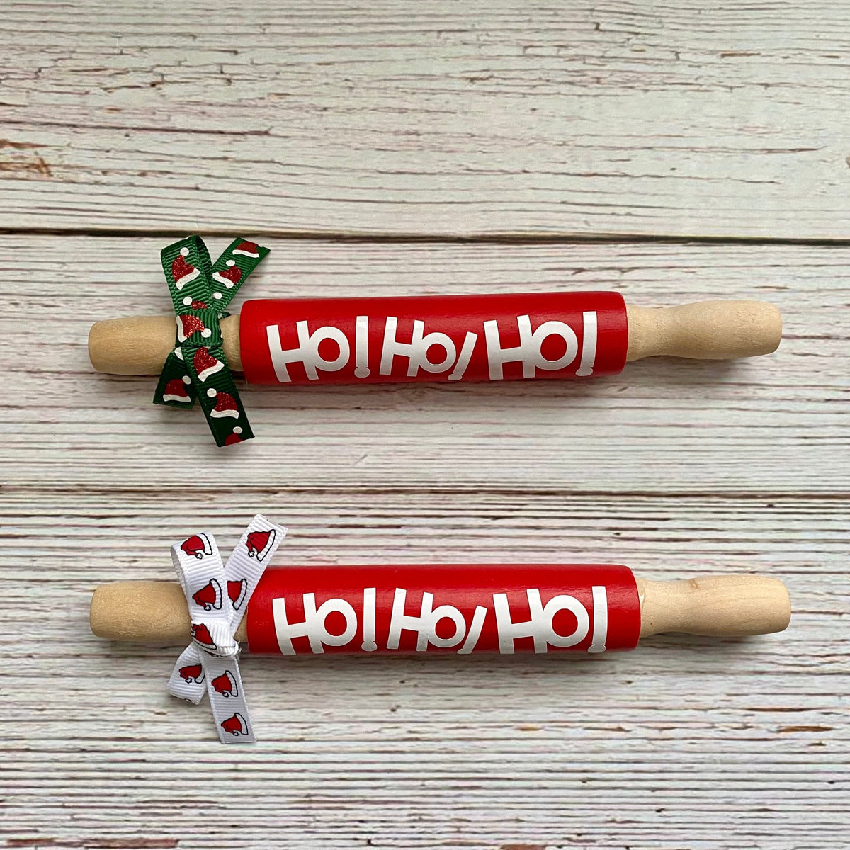 Two wooden rolling pins with red handles printed with 'Ho Ho Ho' in white, tied with festive ribbon bows, displayed on a whitewashed wooden background.