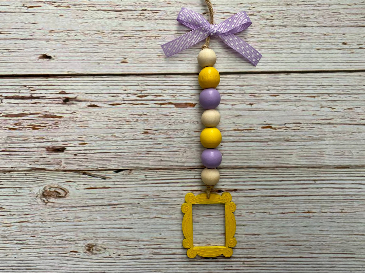 A hanging decorative ornament with a purple patterned ribbon bow at the top, a vertical string of alternating cream, yellow, and lavender beads, and a small yellow picture frame hanging at the bottom, displayed against a rustic whitewashed wood plank background.