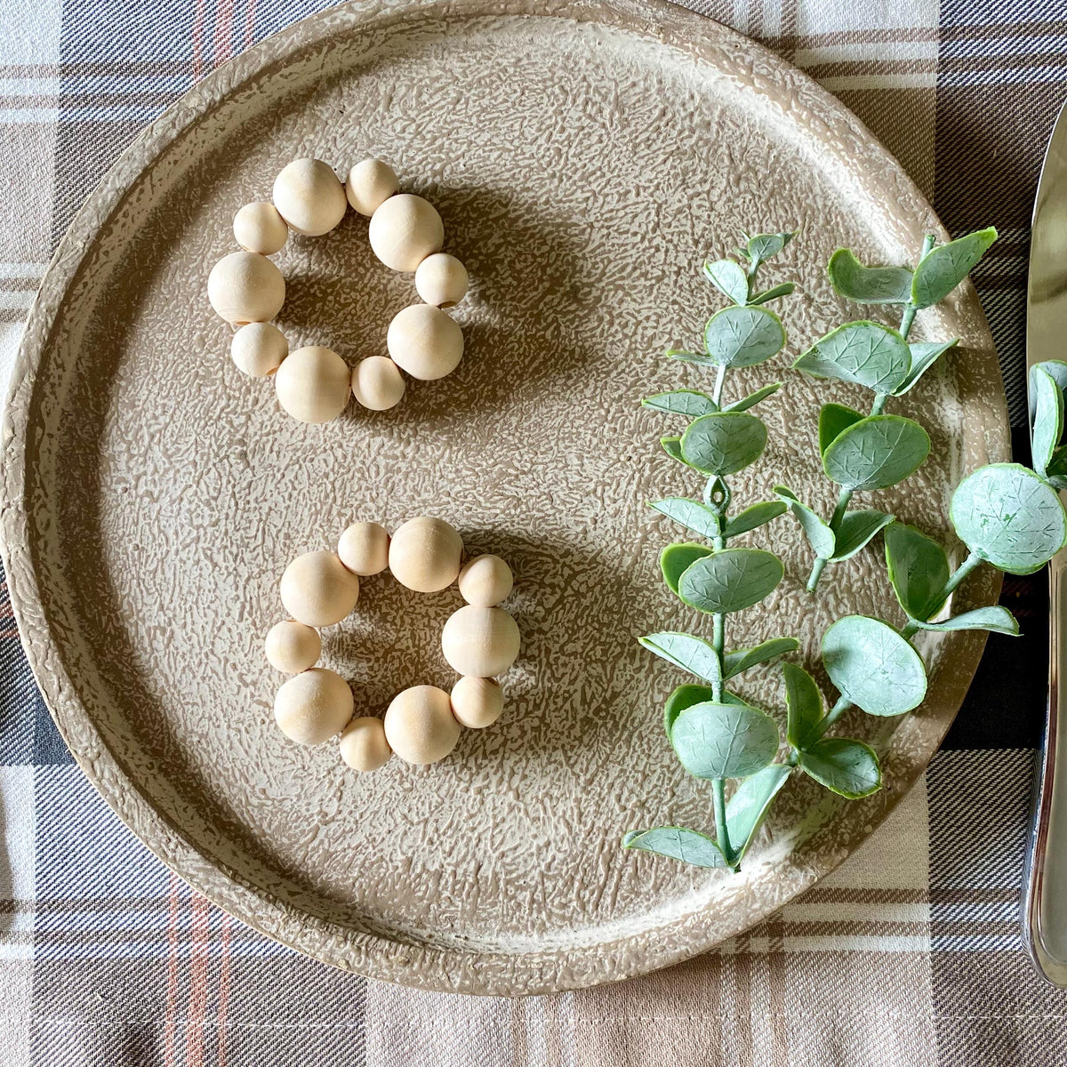 Two circular beaded bracelets made of light natural wood beads are arranged on a textured beige ceramic plate, styled with green eucalyptus leaves on a plaid tablecloth.