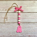 A pink and white wooden ghost ornament hanging from a rustic white wood plank background. The ornament features a pink ghost-shaped wooden cutout at the bottom, a vertical string of pink and white wooden beads, a pink ribbon bow with white polka dots at the top, and a natural jute hanging string.
