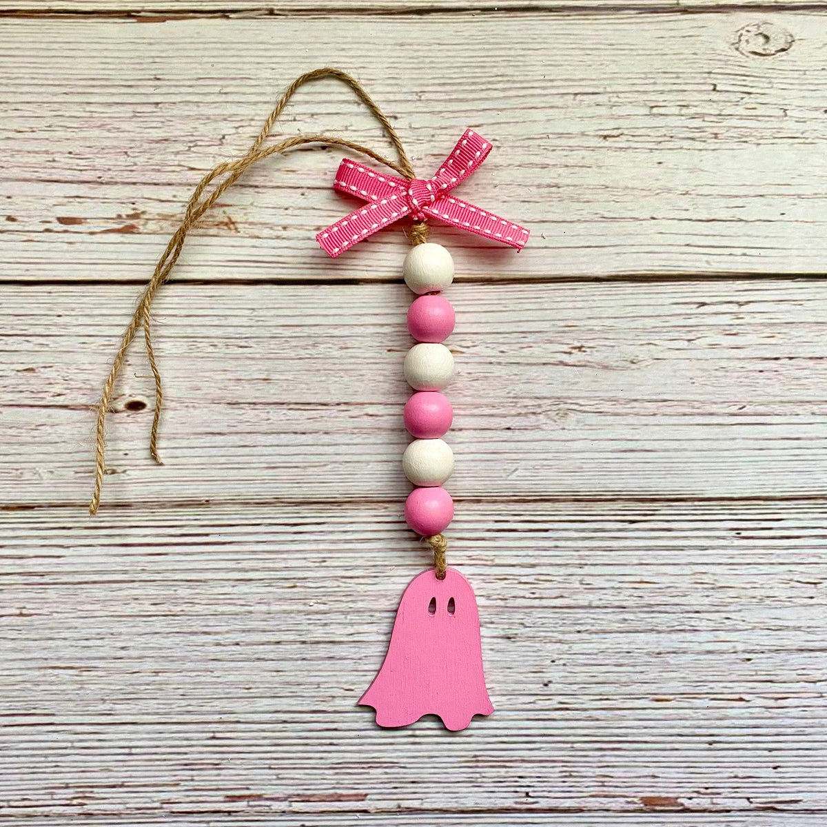 A pink and white wooden ghost ornament hanging from a rustic white wood plank background. The ornament features a pink ghost-shaped wooden cutout at the bottom, a vertical string of pink and white wooden beads, a pink ribbon bow with white polka dots at the top, and a natural jute hanging string.