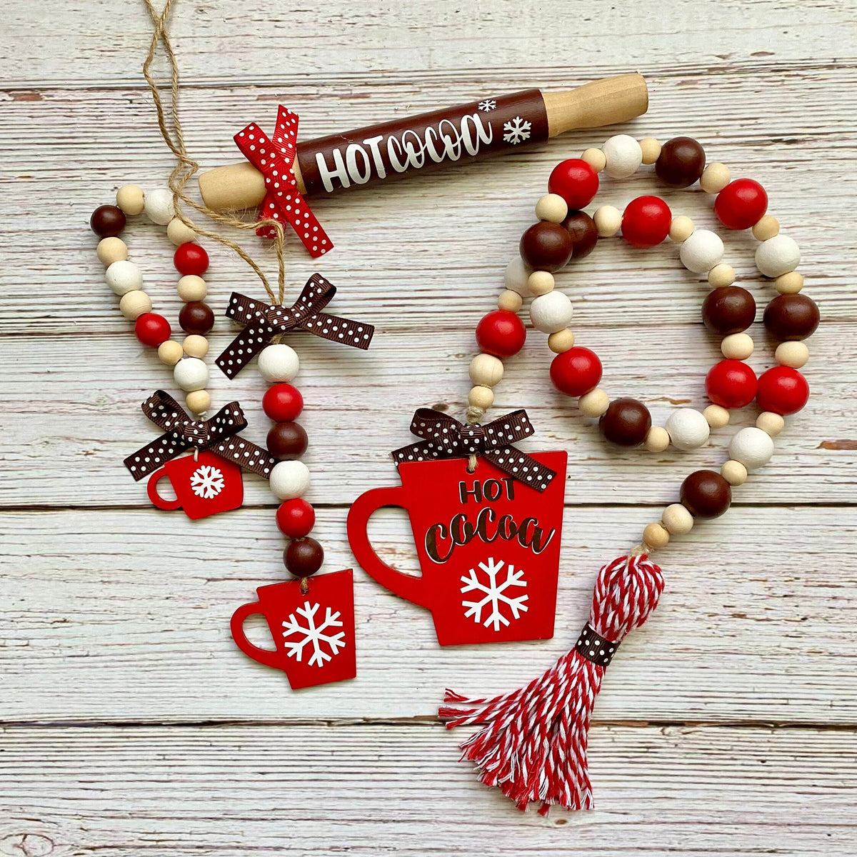 A rustic holiday garland set featuring red and white beaded garlands with a wooden rolling pin ornament, red mug and hot cocoa cutouts, brown polka dot bows, and a red-and-white tassel, arranged on a whitewashed wooden surface.