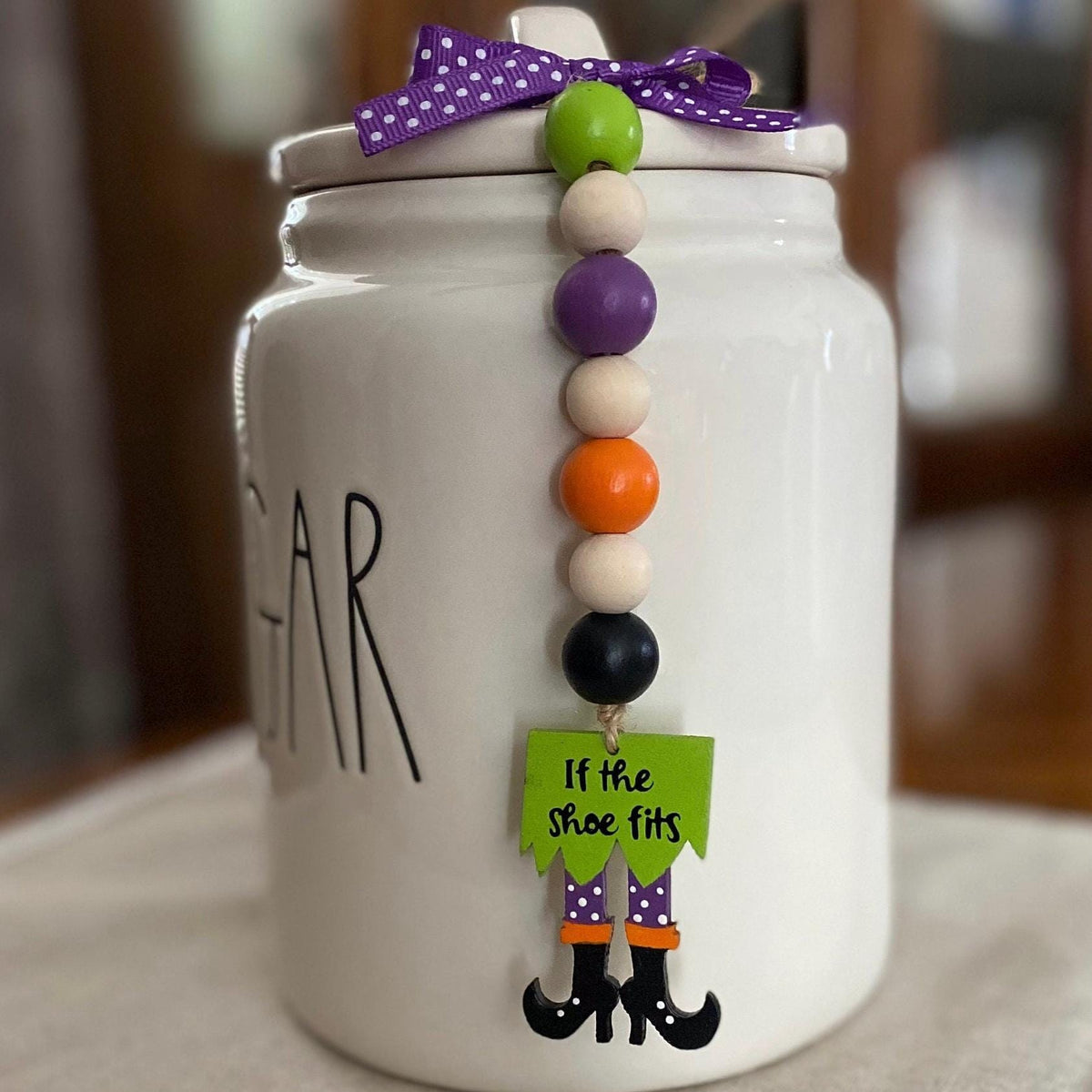 A white ceramic sugar jar with 'SUGAR' written on the front, decorated with a purple polka-dot bow and a hanging beaded garland featuring a witch's legs charm that reads 'If the shoe fits'.