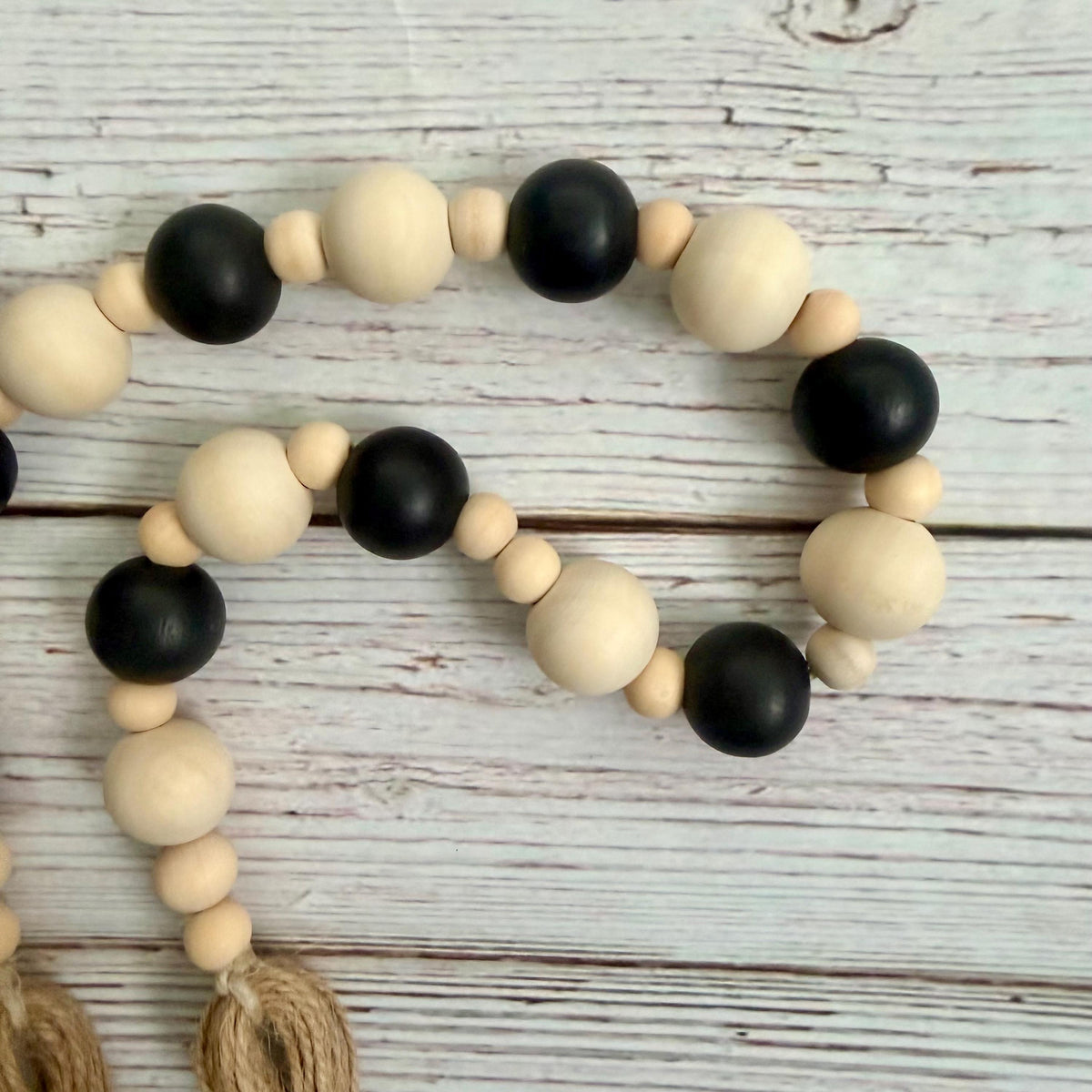 A beaded garland with alternating matte black and natural light wood beads, accented by small tan beads and two knotted jute tassels, displayed on a whitewashed wooden surface.