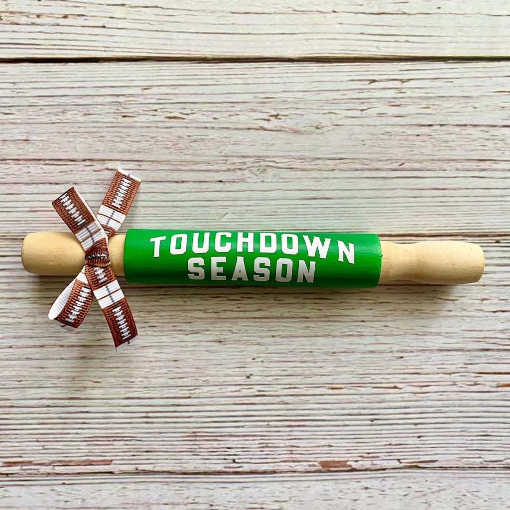 A wooden rolling pin wrapped in bright green that reads 'TOUCHDOWN SEASON' in white letters, tied with a brown and white football-patterned ribbon, on a whitewashed wood plank background.