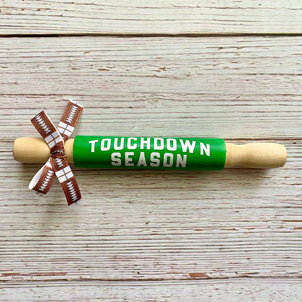 A wooden rolling pin wrapped in bright green that reads 'TOUCHDOWN SEASON' in white letters, tied with a brown and white football-patterned ribbon, on a whitewashed wood plank background.