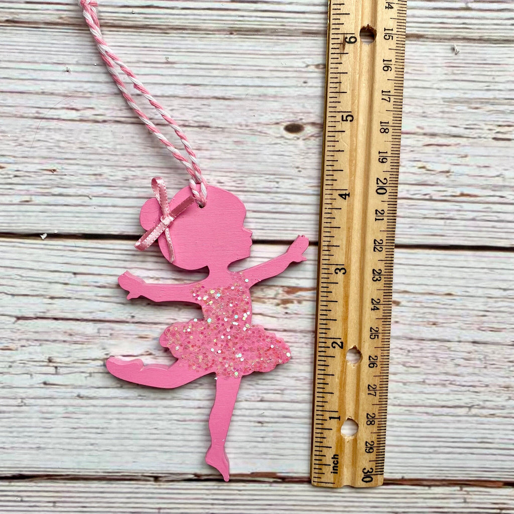 A pink glittery ballerina silhouette ornament with a pink and white striped hanging string, displayed next to a wooden ruler on a whitewashed wood background.
