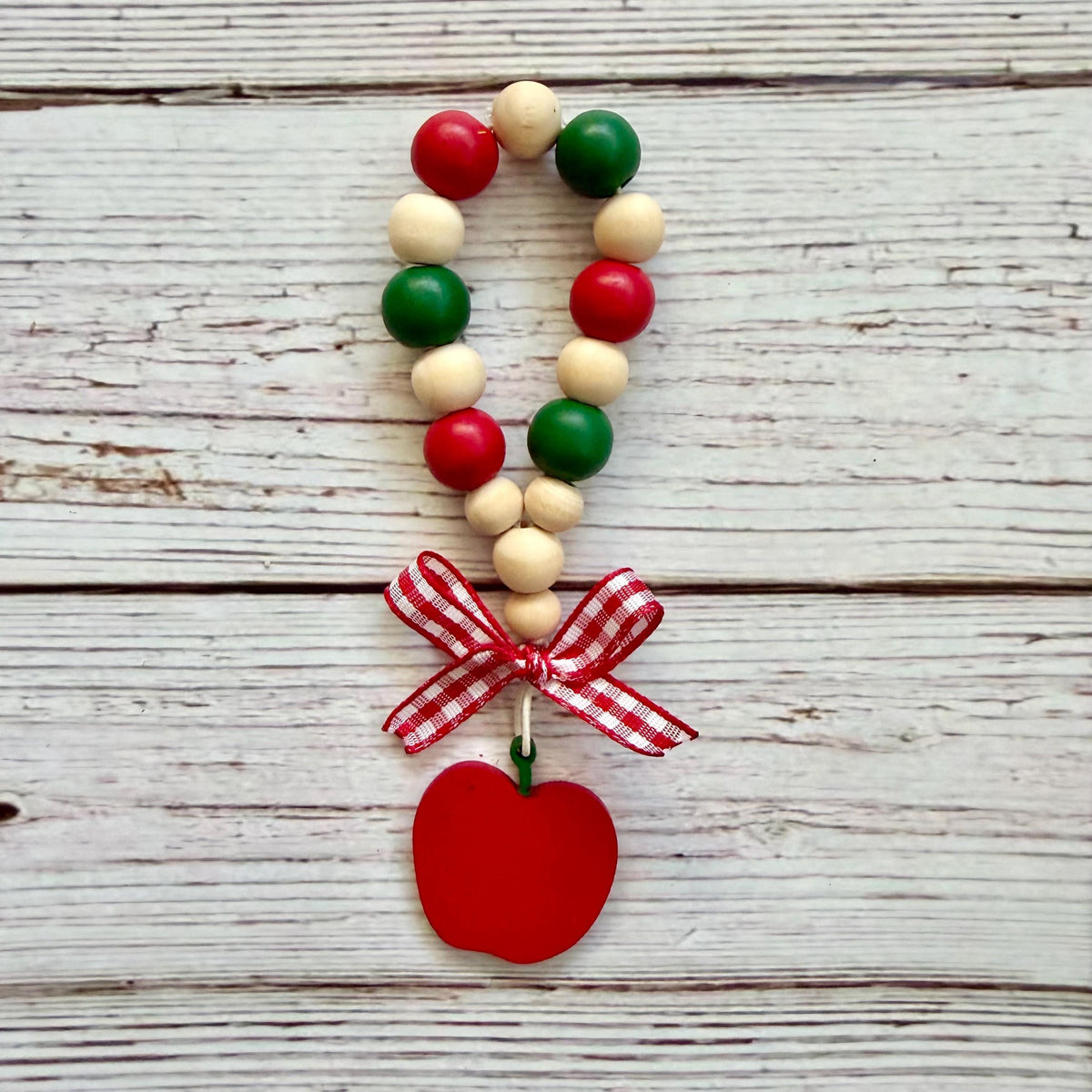A beaded loop ornament with alternating red, green, and natural wood beads, finished with a red-and-white gingham ribbon bow and a red apple-shaped charm, displayed on a whitewashed wooden surface.