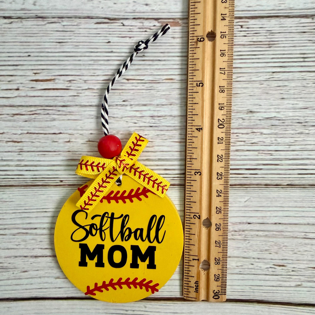 A round yellow 'Softball MOM' ornament styled like a softball with red stitching, a black-and-white hanging string, and a red bead, shown next to a wooden ruler on a whitewashed wood background.