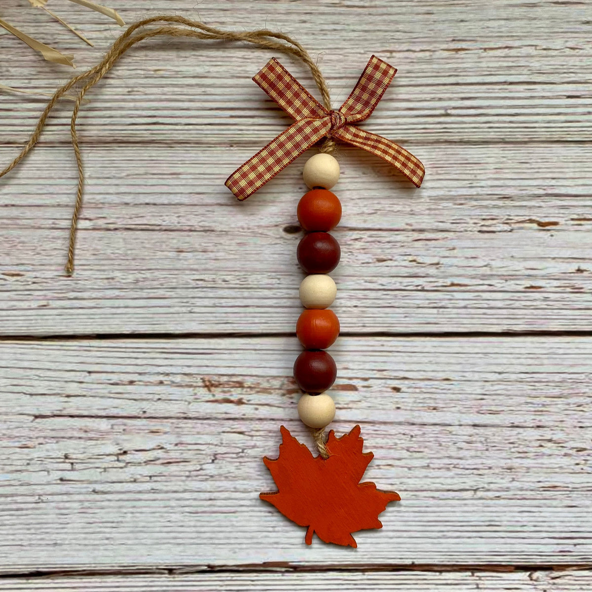 Autumn-themed wooden bead garland ornament with a gingham ribbon bow, colorful wooden beads, and a red maple leaf charm on a distressed white wood background.