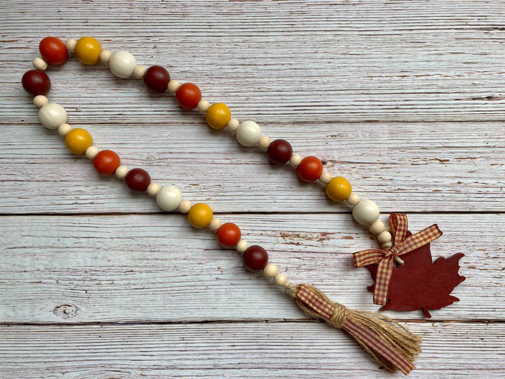 Autumn-themed wooden bead garland with orange, red, yellow, and cream beads, finished with a tassel and a red maple leaf charm, displayed on a rustic white-washed wooden background.