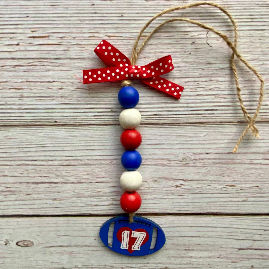 A patriotic beaded ornament featuring red, white, and blue wooden beads, a red polka-dot ribbon bow, and a blue football-shaped tag with the number 17, hanging against a rustic whitewashed wood plank background.