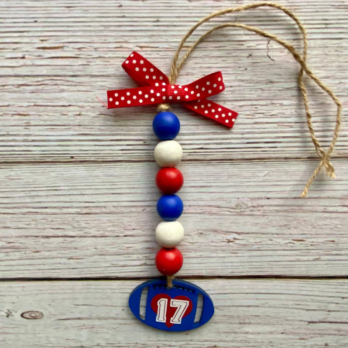 A patriotic beaded ornament featuring red, white, and blue wooden beads, a red polka-dot ribbon bow, and a blue football-shaped tag with the number 17, hanging against a rustic whitewashed wood plank background.