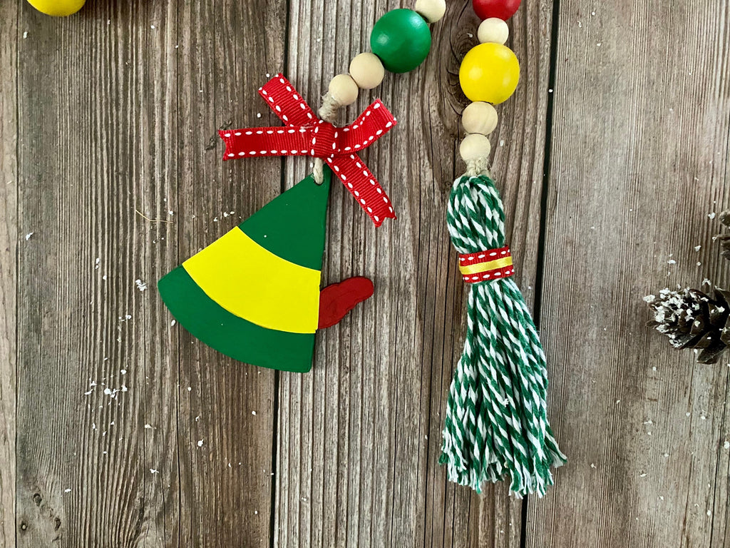 A Christmas ornament made from a painted Christmas elf hat with a red ribbon bow, hanging next to a green and white striped tassel bead garland on a rustic wooden background.