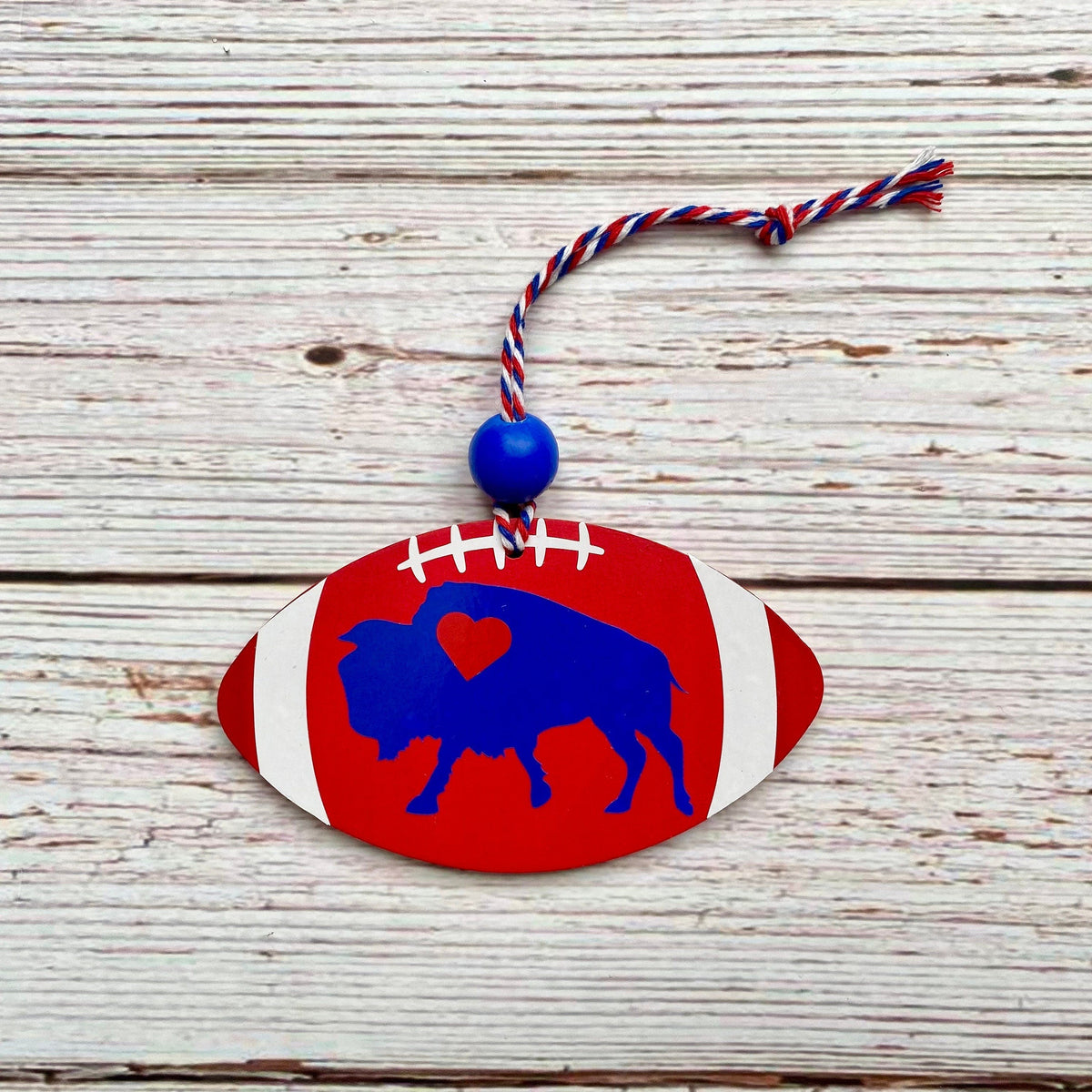 A patriotic football-shaped ornament featuring a blue buffalo silhouette with a red heart on its chest, hanging from a red, white, and blue string on a whitewashed wooden background.