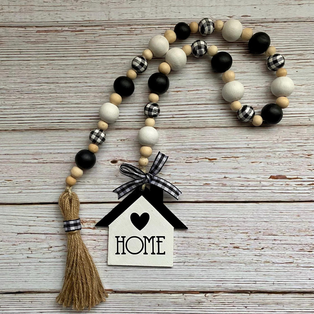 A rustic wooden bead garland with black, white, and gingham pattern beads, a jute tassel, and a house-shaped 'HOME' ornament, displayed on a whitewashed wood background.