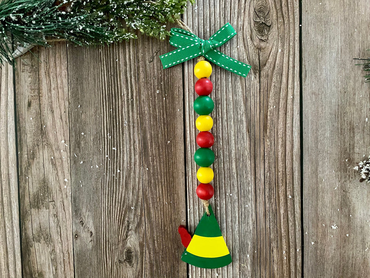 A Christmas-themed hanging ornament featuring a green ribbon bow at the top, a vertical string of alternating red and yellow wooden beads, and an elf hat wood charm in green and yellow, displayed against a rustic wooden plank background.