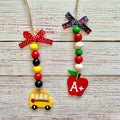 Two personalized wooden bead garlands, one shaped like a yellow school bus and the other like a red apple with an A+ sign, hanging on a rustic white wood background.