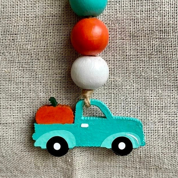 A handmade beaded garland featuring a turquoise painted pickup truck carrying an orange pumpkin, suspended from a string of wooden beads in turquoise, orange, white, and turquoise, laid flat on a beige linen background.