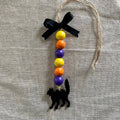 A Halloween hanging ornament made from a natural jute twine loop with a black ribbon bow, colorful wooden beads in yellow, orange, and purple, and a black silhouette of a cat at the bottom, displayed on a textured beige linen fabric background.