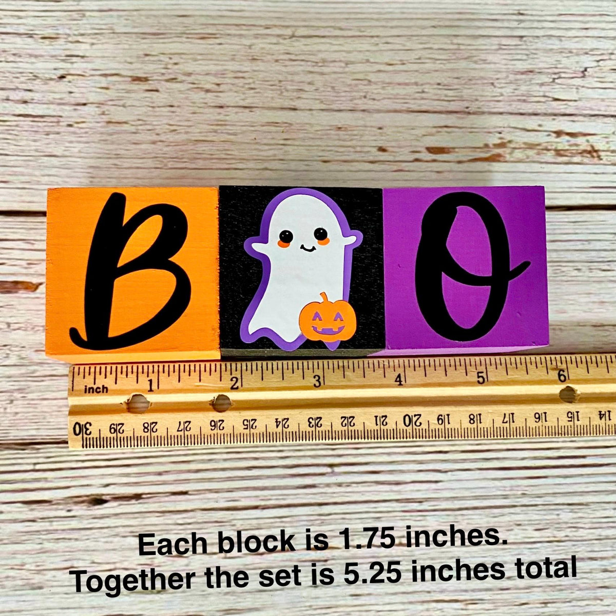 Three Halloween-themed wooden blocks spelling BOO, shown with a ruler for scale on a whitewashed wood background. The left block is orange with a black letter B, the middle block is black with a white ghost holding an orange pumpkin, and the right block is purple with a black letter O.