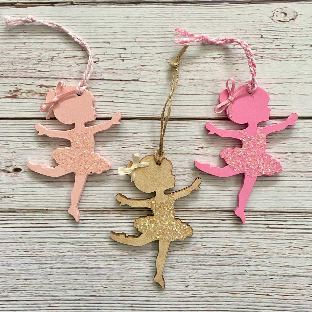 Three ballerina-shaped hanging ornaments on a whitewashed wood background. One ornament is light pink with glitter, one is bright pink with glitter, and one is natural wood with glitter.