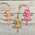 Three ballerina-shaped hanging ornaments on a whitewashed wood background. One ornament is light pink with glitter, one is bright pink with glitter, and one is natural wood with glitter.
