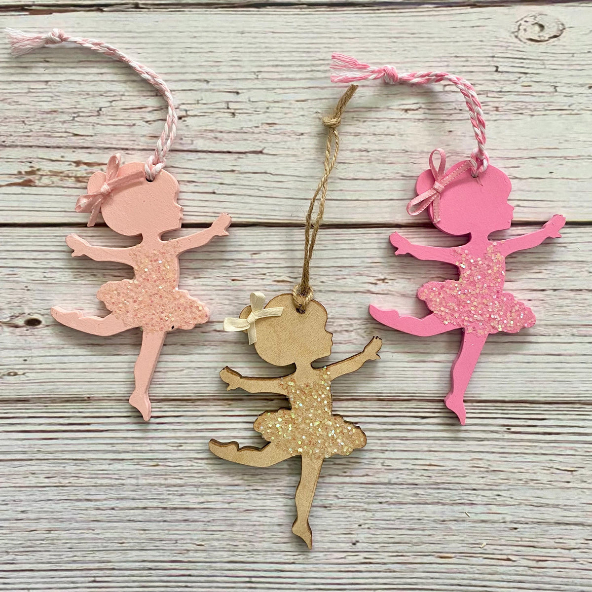 Three ballerina-shaped hanging ornaments on a whitewashed wood background. One ornament is light pink with glitter, one is bright pink with glitter, and one is natural wood with glitter.