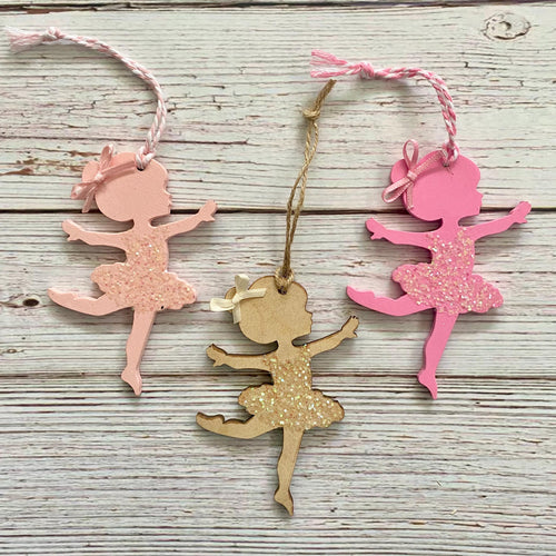 Three ballerina-shaped hanging ornaments on a whitewashed wood background. One ornament is light pink with glitter, one is bright pink with glitter, and one is natural wood with glitter.