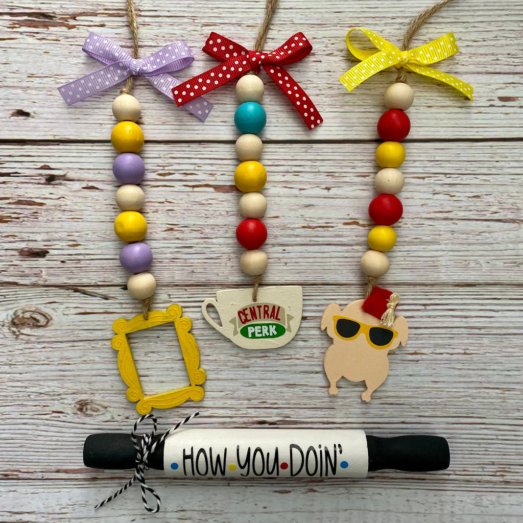 Three colorful beaded Christmas tree ornaments with red, yellow, purple, and teal beads hang on a rustic white-washed wooden background. The ornaments feature various coffee-themed charms, including a 'Central Perk' mug, a 'How You Doin'' sign, a coffee cup, and a dog wearing sunglasses.