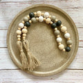 A beaded garland with two tassels, featuring natural wood beads, sage green beads, and dark charcoal beads, displayed on a round textured ceramic plate over a white wood plank background.