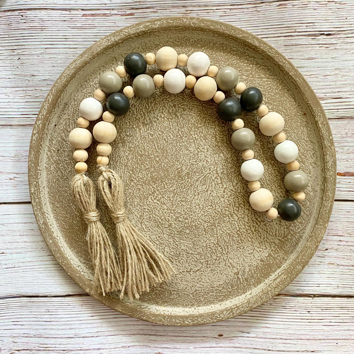 A beaded garland with two tassels, featuring natural wood beads, sage green beads, and dark charcoal beads, displayed on a round textured ceramic plate over a white wood plank background.