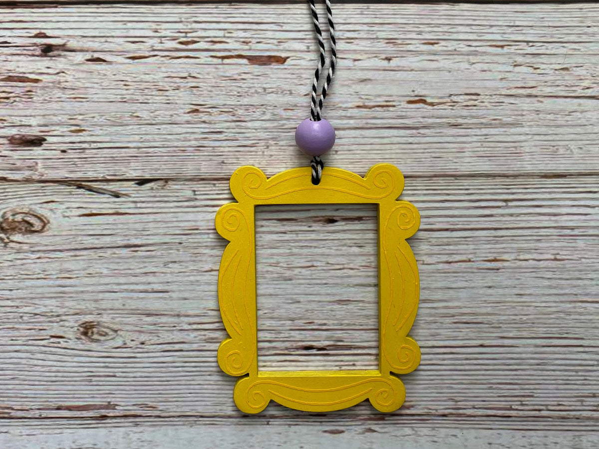 A yellow picture frame ornament with a lavender purple bead and a black-and-white twisted cord, displayed on a rustic white-washed wooden background.