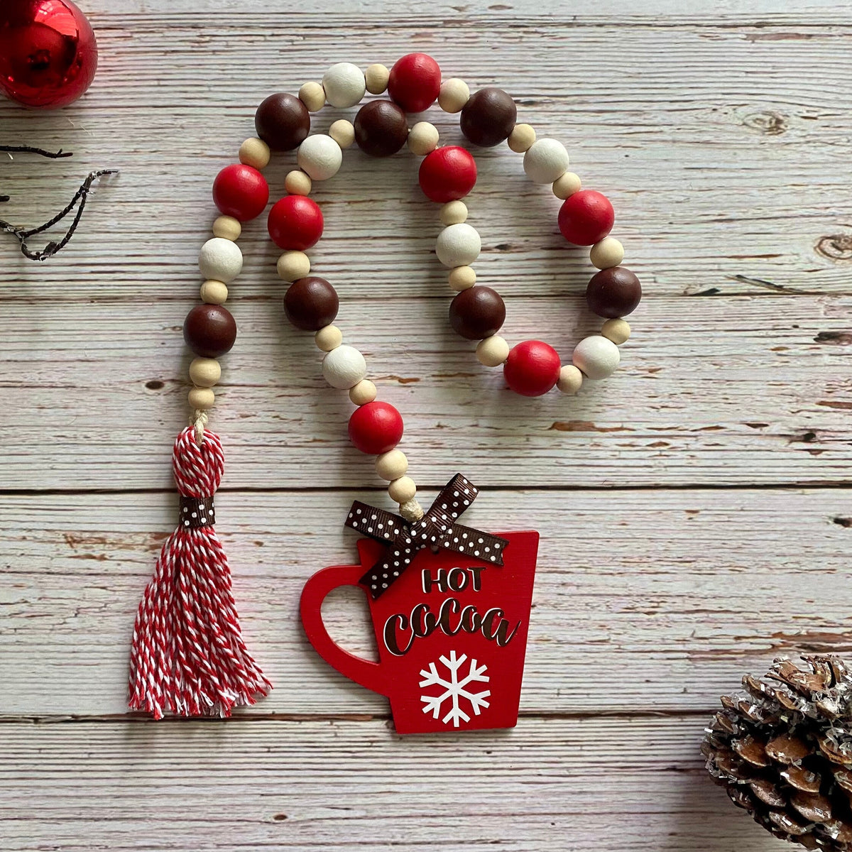 A Christmas-themed beaded garland with red, white, and dark brown wooden beads, a red and white striped tassel, and a red mug charm that reads 'Hot Cocoa' with a white snowflake. The garland is displayed on a rustic white wood background with holiday decorations including red ornaments and a pinecone.